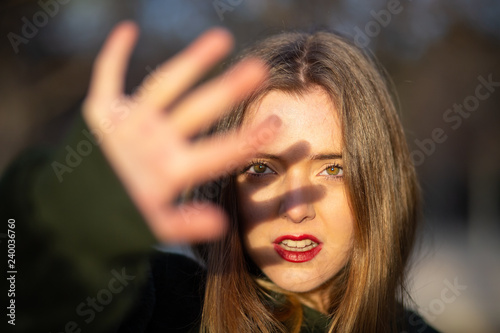 Lovely young female with bright makeup trying to cover face with hand from sunlight while standing on blurred background of park