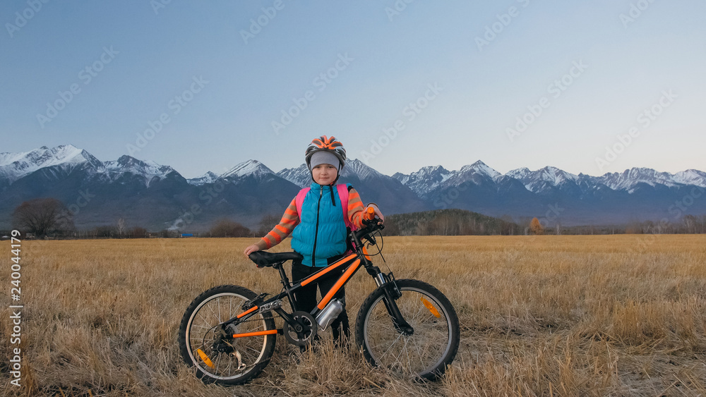 Obraz premium One caucasian children walk with bike in wheat field. Little girl walking black orange cycle on background of beautiful snowy mountains. Biker stand with backpack and helmet.