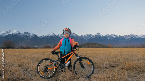 One caucasian children walk with bike in wheat field. Little girl walking black orange cycle on background of beautiful snowy mountains. Biker stand with backpack and helmet.