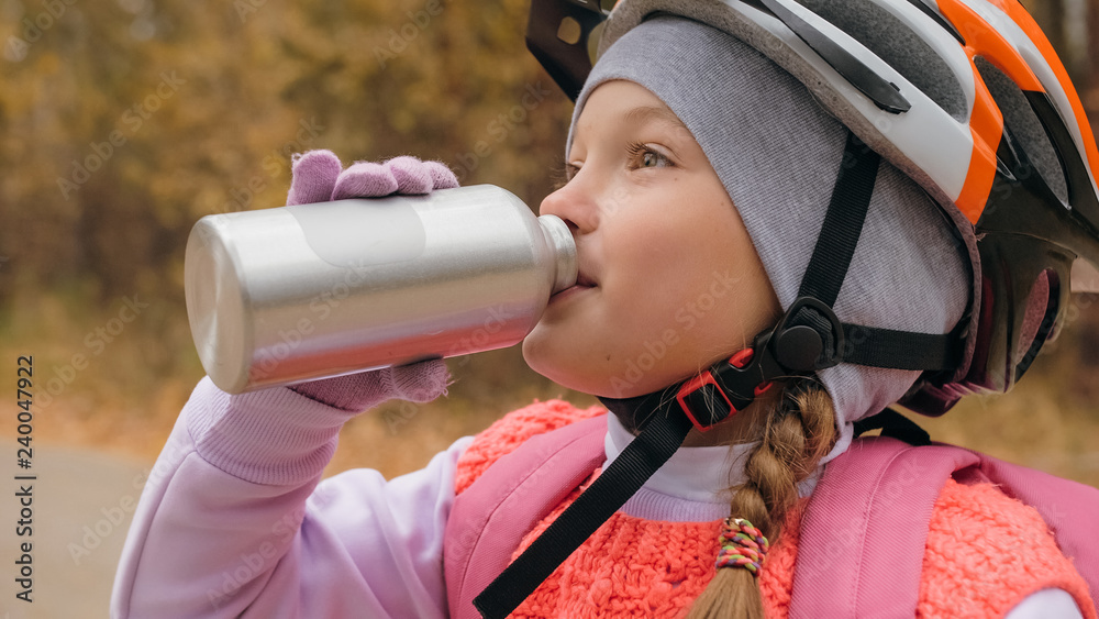 Fototapeta premium Kid drink water from an aluminum flask bottle. One caucasian children rides bike road in autumn park. Little girl riding black orange mtb cycle in forest. Biker motion ride with backpack and helmet.
