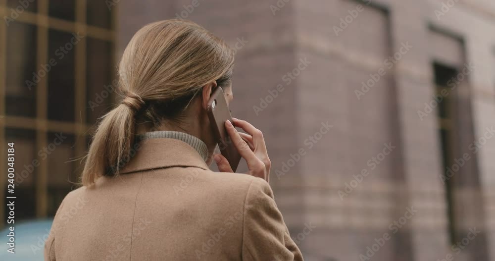 Back view on the Caucasian woman in a coat walking the street and speaking on the mobile phone. Close up. Rear. Outside. Back view on the Caucasian woman in a coat walking the street and speaking on the mobile phone. Close up. Rear. Outside.