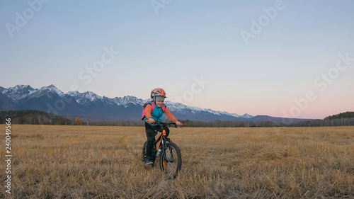 One caucasian children rides bike in wheat field. Little girl riding black orange cycle on background of beautiful snowy mountains. Biker motion ride with backpack, helmet. Mountain bike hardtail.