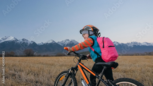 One caucasian children walk with bike in wheat field. Little girl walking black orange cycle on background of beautiful snowy mountains. Biker stand with backpack and helmet.