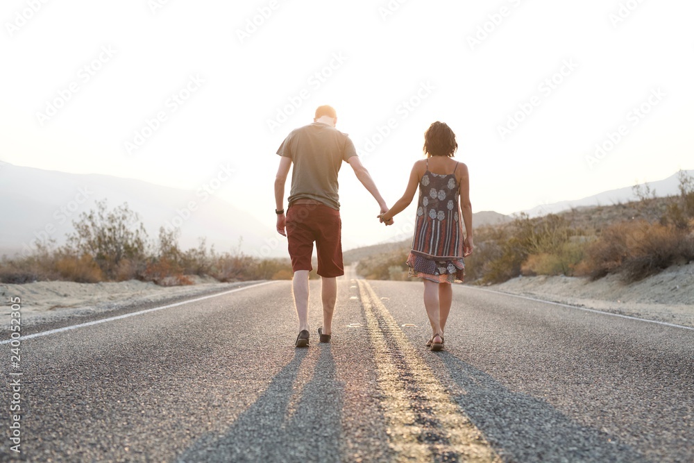 Young man and woman holding hands walking down desert road during sunrise Young man and woman holding hands walking down desert road during sunrise