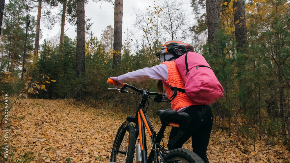 Fototapeta premium One caucasian children walk with bike in autumn park. Little girl walking black orange cycle in forest. Kid goes do bicycle sports. Biker motion ride with backpack and helmet. Mountain bike hardtail.