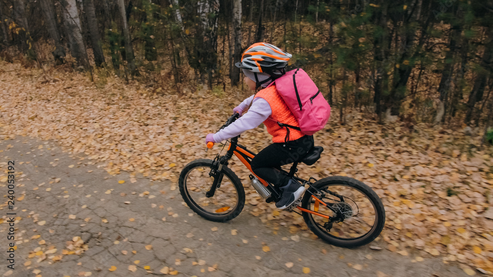 One caucasian children rides bike road in autumn park. Little girl riding black orange cycle in forest. Kid goes do bicycle sports. Biker motion ride with backpack and helmet. Mountain bike hardtail. One caucasian children rides bike road in autumn park. Little girl riding black orange cycle in forest. Kid goes do bicycle sports. Biker motion ride with backpack and helmet. Mountain bike hardtail.
