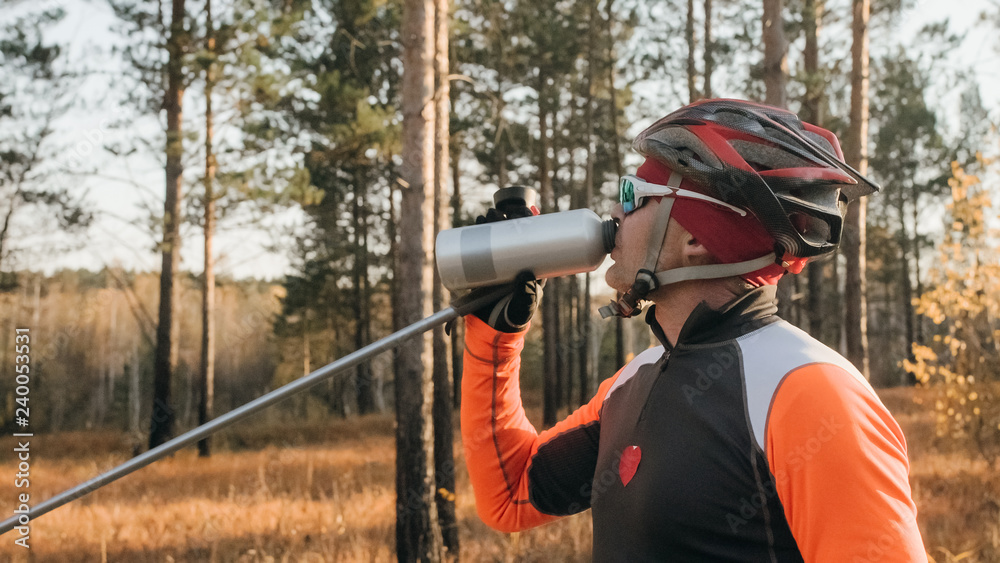 Training an athlete on the roller skaters. Biathlon ride on the roller skis with ski poles, in the helmet. Autumn workout. Roller sport. Adult man riding on skates. Athlete drinks water from flask.