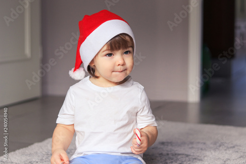Cute little baby boy in red Santa hat holding Christmas striped candy cane in his hand. Happy careless childhood. Childrend and sweets concept. Mock up. photo