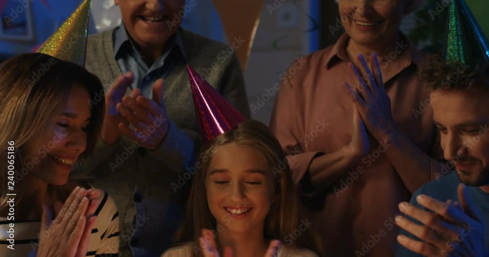Birthday teen smiled girl surrounding by the whole family and blowing candles on the cake in the dark room. Indoor.
