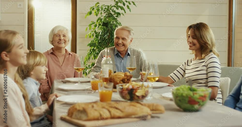 Caucasian family of grandparents, parents and kids sitting at the table and having dinner together in the living room. Indoor. Caucasian family of grandparents, parents and kids sitting at the table and having dinner together in the living room. Indoor.