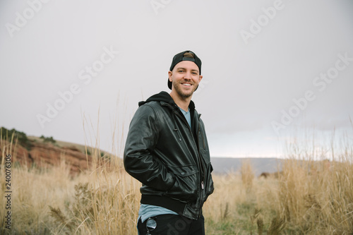 Portrait of Young Good Looking Handsome Man Smiling with Backwards Hat in Leather Jacket Outside in Isolated Rural Field of Tall Grass in the Meadow Nature Outside Autumn Season © MeganMahoneyPhotos