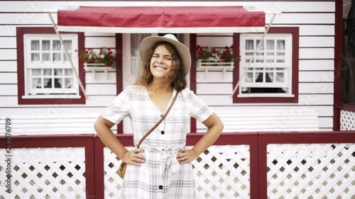 Lovely young woman in checkered dress and hat smiling and keeping hands on waist while standing near fence of cute building on street of Costa Nova, Portugal