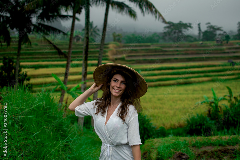 Beautiful young woman in shine through dress touch asian, Vietnam rice hat. Girl walk at typical Asian hillside with rice farming, in cascad rice field with palm, Bali, Indonesia.