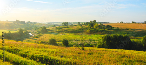 Sunny country summer landscape with green meadows hills golden wheat fields and distant woods at sunrise.