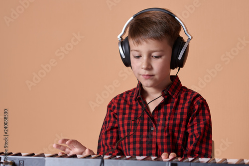 The portrait of a good-looking European boy on a peach background. He is making his first steps in music. He is playing electric piano and using headphones.