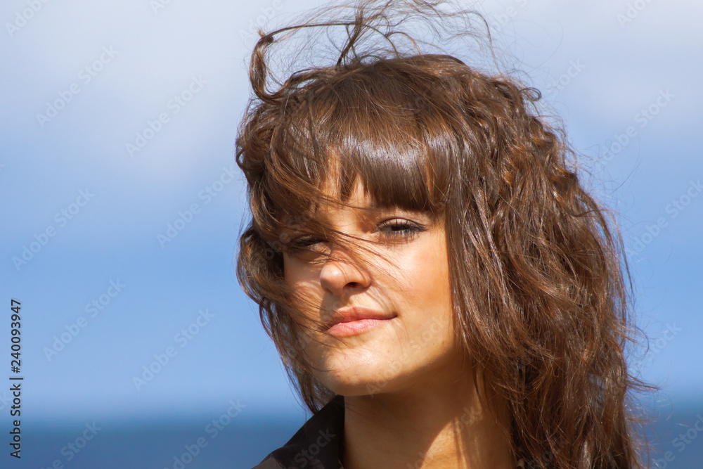 closeup portrait of beautiful smiling woman in black jacket, with long dark brown curly hair outdoor at sunny windy day, on blue sky background looking into the camera closeup portrait of beautiful smiling woman in black jacket, with long dark brown curly hair outdoor at sunny windy day, on blue sky background looking into the camera