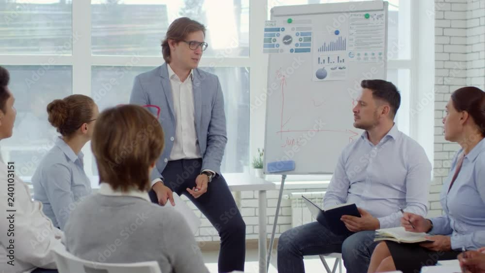 Professional business people sitting in circle on chairs, taking notes and discussing ideas with male executive in office room with flipchart