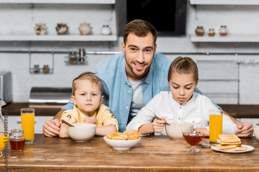 smiling father looking at camera and embracing siblings eating porridge at table smiling father looking at camera and embracing siblings eating porridge at table