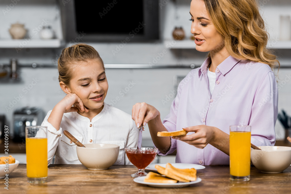 smiling girl looking at pretty woman spreading jam on toast in kitchen smiling girl looking at pretty woman spreading jam on toast in kitchen