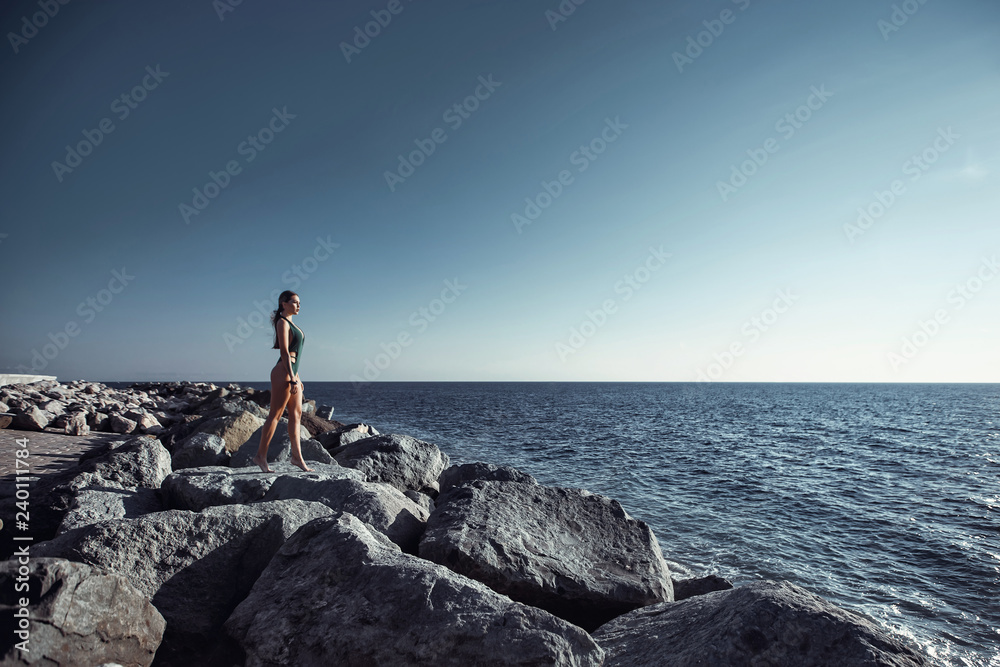 Beautiful girl in a swimsuit standing on a rock by the ocean. Island, sunset, rest, sun, travel, freedom.