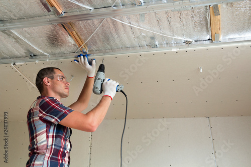 Young man in goggles fixing drywall suspended ceiling to metal frame using electrical screwdriver on ceiling insulated with shiny aluminum foil. Renovation, construction, do it yourself concept. photo