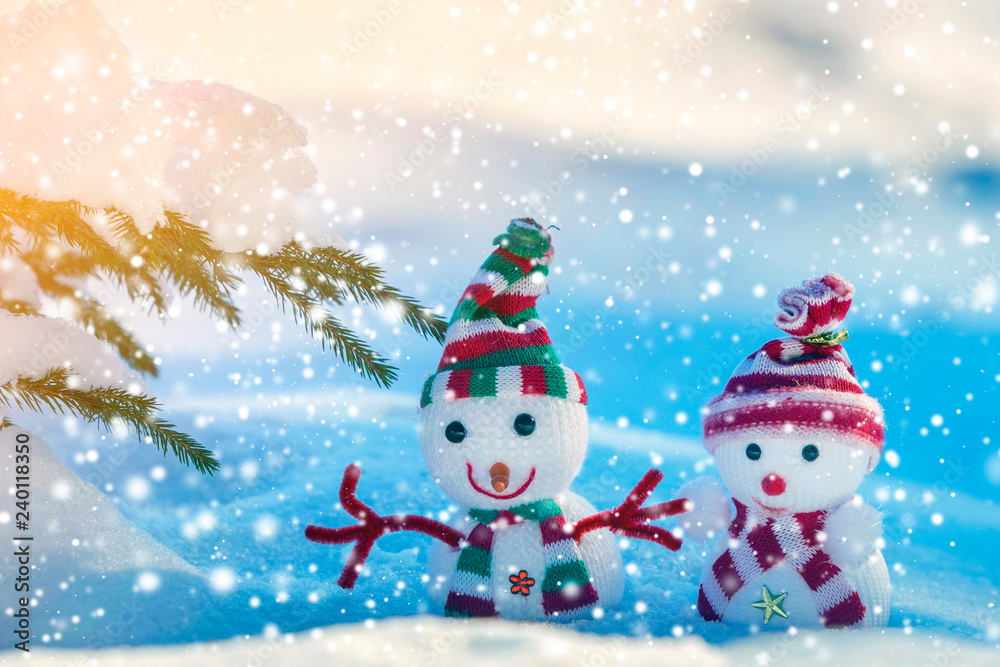 Fototapeta premium Two small funny toys baby snowman in knitted hats and scarves in deep snow outdoors on bright blue and white copy space background. Happy New Year and Merry Christmas greeting card.