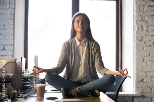 Serene calm business woman sit on office desk taking break for meditation, mindful employee doing yoga exercise in lotus pose for relaxation at workplace, no stress relief, balance at work concept