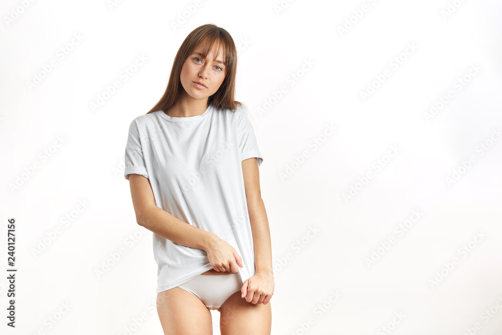 Foto de Attractive young woman in oversized white t-shirt, being in  underwear, shows perfect slim body and healthy skin, posing over white  studio background. do Stock