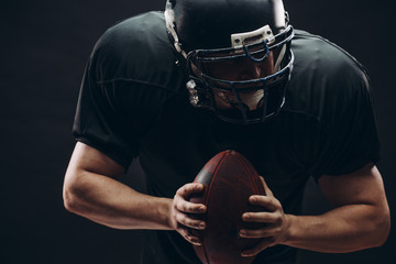 American football player with black helmet and armour running in motion, holding ball, getting ready to score a goal, close up shot over dark background