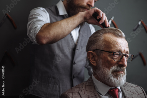 Barber making male haircut. Hairdresser cutting hair of his rich client, dressed in expensive stylish man s suit of clothes and holding a glass of alcoholic beverage