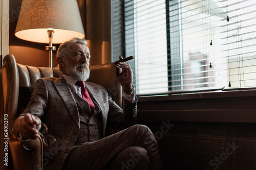 Portrait of experienced unshaven grey-haired pensioner smoking cigar while sitting in arm-chair in apartment. Age, Confidence and prosperity concept