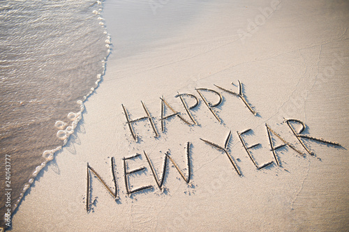 Happy New Year message handwritten in smooth, clean sand with a fresh wave coming up the beach