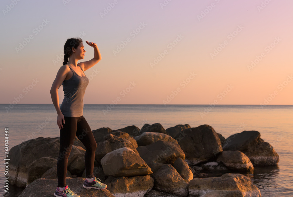 sporty girl standing on a rock against the sea with a smile looking at the sunset