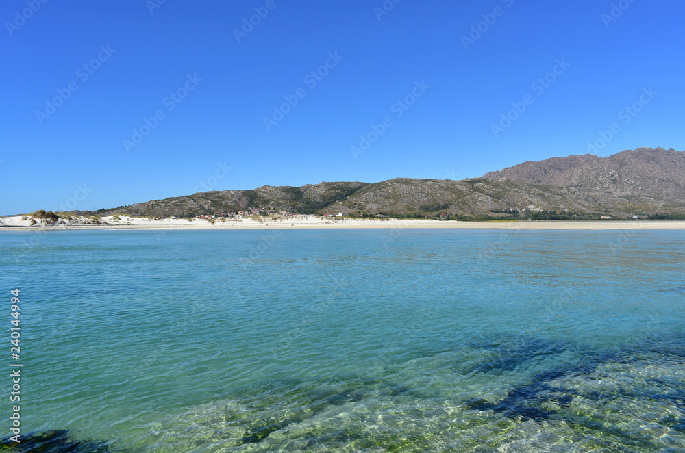 Beach with clear and turquoise water and small mountain. Sunny day, blue sky. Galicia, Spain.
