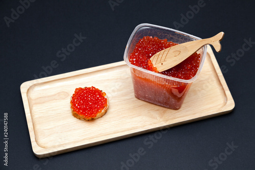 An open plastic jar of red salmon caviar with a fish-shaped spoon and a gourmet snack round sandwich on a rectangular wooden plate on a black background. Concept useful delicacy, seafood diet photo