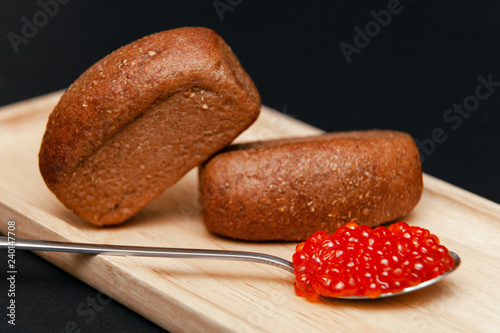 Closeup macro two small pieces black bread with cumin and metal spoon with slide of red salmon caviar gourmet snack on rectangular wooden plate on black background. Concept healthy seafood appetizer photo