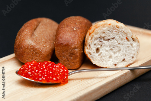 Closeup small pieces black cumin and white grain bread, metal spoon with slide of red salmon caviar on rectangular wooden plate on black background. Concept gourmet healthy seafood appetizer photo