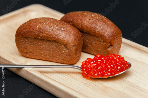 Closeup macro two small pieces black bread with cumin and metal spoon with slide of red salmon caviar gourmet snack on rectangular wooden plate on black background. Concept healthy seafood appetizer photo