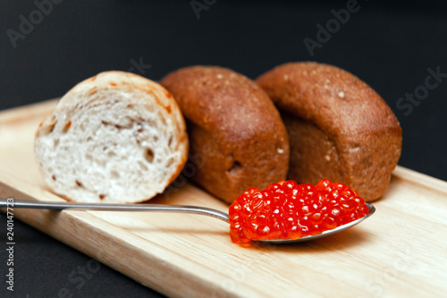 Closeup small pieces black cumin and white grain bread, metal spoon with slide of red salmon caviar on rectangular wooden plate on black background. Concept gourmet healthy seafood appetizer photo