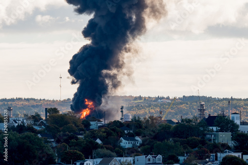 Large fire at an oil refinery seen from a distance. Only parts of the refinery visible behind the trees. Thick black smoke rises from the flames.