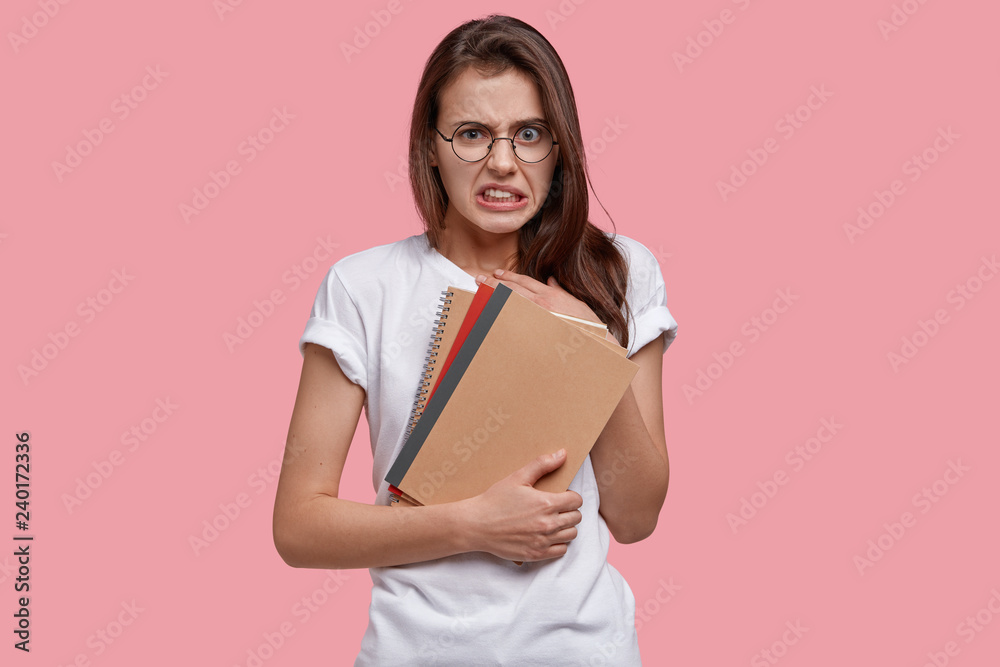 Fototapeta premium Displeased college student lady has unhappy facial expression, clenches teeth, carries spiral notebooks, has no inspiration for work, isolated over pink studio wall. Youth and studying concept