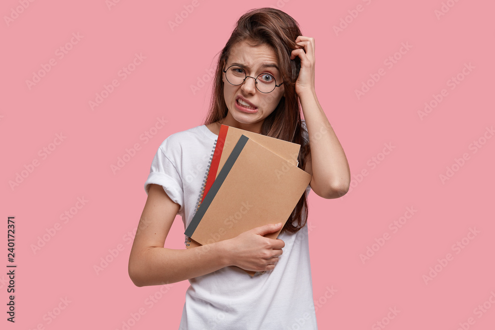 Displeased young woman scratches head, clenches teeth, looks in bewilderment, thinks about creative ideas for writing essay, holds notepads, dressed in casual clothes, isolated on pink background Displeased young woman scratches head, clenches teeth, looks in bewilderment, thinks about creative ideas for writing essay, holds notepads, dressed in casual clothes, isolated on pink background