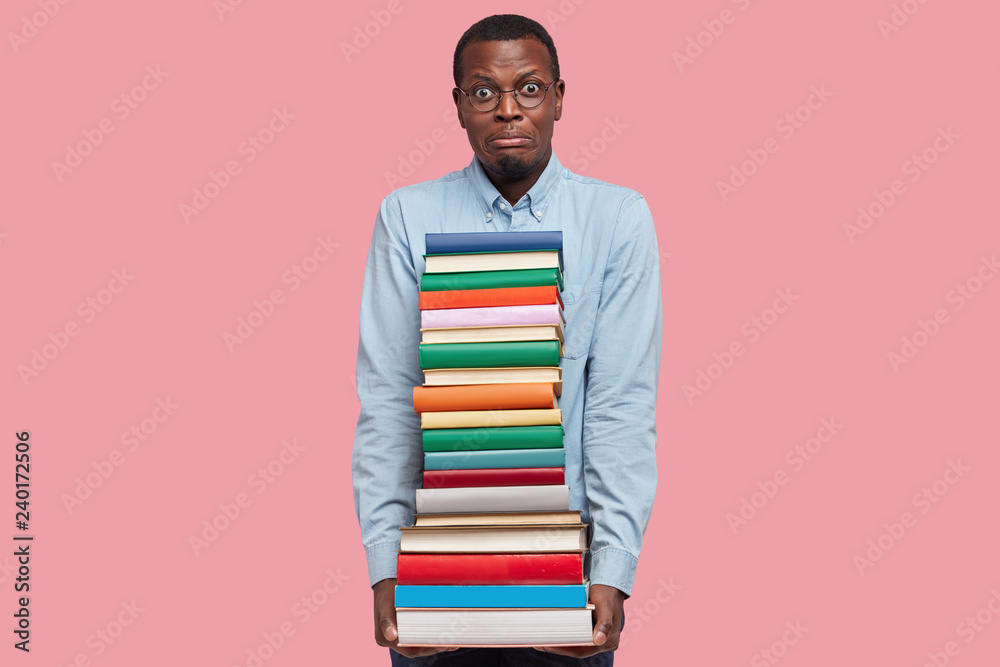 Shot of puzzled dissatisfied dark skinned man holds heavy pile of books, dressed in formal shirt, models over pink studio wall, busy with studying scientific literature, isolated over pink studio wall Shot of puzzled dissatisfied dark skinned man holds heavy pile of books, dressed in formal shirt, models over pink studio wall, busy with studying scientific literature, isolated over pink studio wall