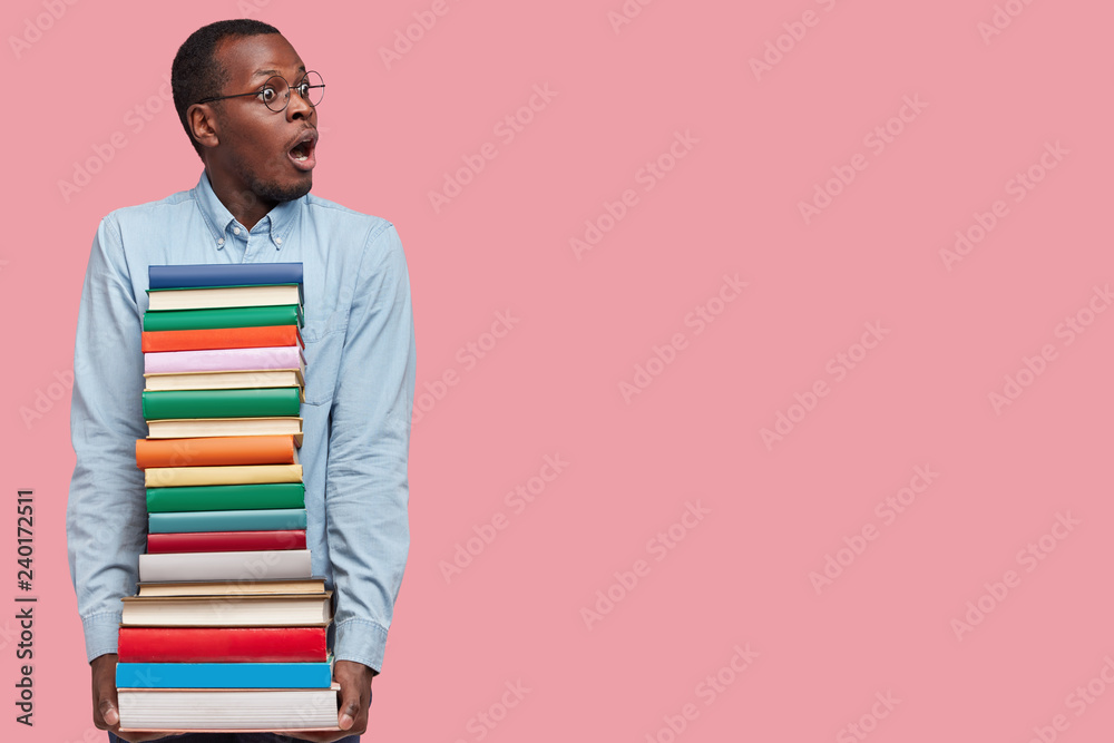 Studio shot of stupefied dark skinned black male professor, holds many books neatly arranged, prepares for seminar or conducting lecture, dressed formally, isolated on pink wall with copy space Studio shot of stupefied dark skinned black male professor, holds many books neatly arranged, prepares for seminar or conducting lecture, dressed formally, isolated on pink wall with copy space