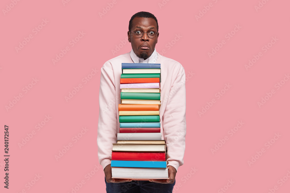Horizontal shot of African American student has puzzled expression, surprised look, holds thick encyclopedia and scientific literature, wears round spectacles, isolated over pink studio wall Horizontal shot of African American student has puzzled expression, surprised look, holds thick encyclopedia and scientific literature, wears round spectacles, isolated over pink studio wall
