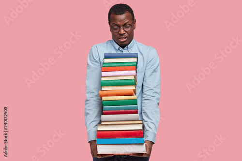 Photography Indoor shot of displeased African American scientist or student carries heavy pile of textbooks, wears elegant shirt, models over pink studio wall, realizes how much he should learn