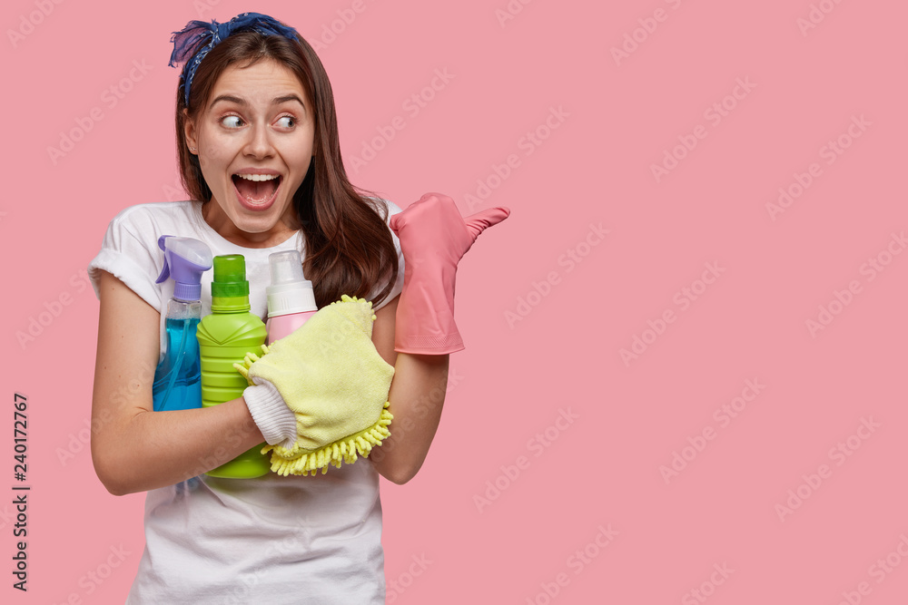 Glad Caucasian woman with positive expression, wears rubber gloves, points aside with thumb, holds spray and detergent in hands, dressed in casual white t shirt, models over pink studio wall Glad Caucasian woman with positive expression, wears rubber gloves, points aside with thumb, holds spray and detergent in hands, dressed in casual white t shirt, models over pink studio wall