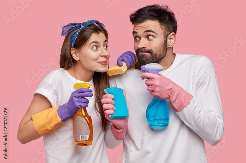 Funny European wife and husband hold mop and bottle of spray, wear rubber protective gloves, look positively at each other, isolated over pink background. Chambermaid works about house at home photo