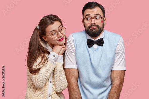 Comic young woman with two pony tails looks with love at boyfriend who has red lipstick on cheek after recieving kiss, feel awkward as have first date, wear elegant clothes and big spectacles