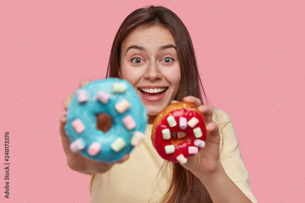 Happy glad lady with toothy smile holds delicious doughnuts, looks joyfully at camera, suggests to taste it, isolated over pink background. Delighted female model cooked tasty donuts on Christmas Happy glad lady with toothy smile holds delicious doughnuts, looks joyfully at camera, suggests to taste it, isolated over pink background. Delighted female model cooked tasty donuts on Christmas